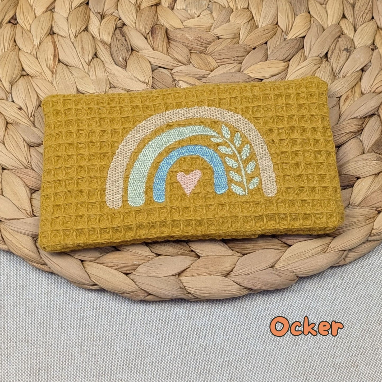 Auf dem Bild ist eine Stoffhülle für ein Kühlakku in der Stofffarbe beere zu sehen. Mittig ist ein Regenbogen gestickt.