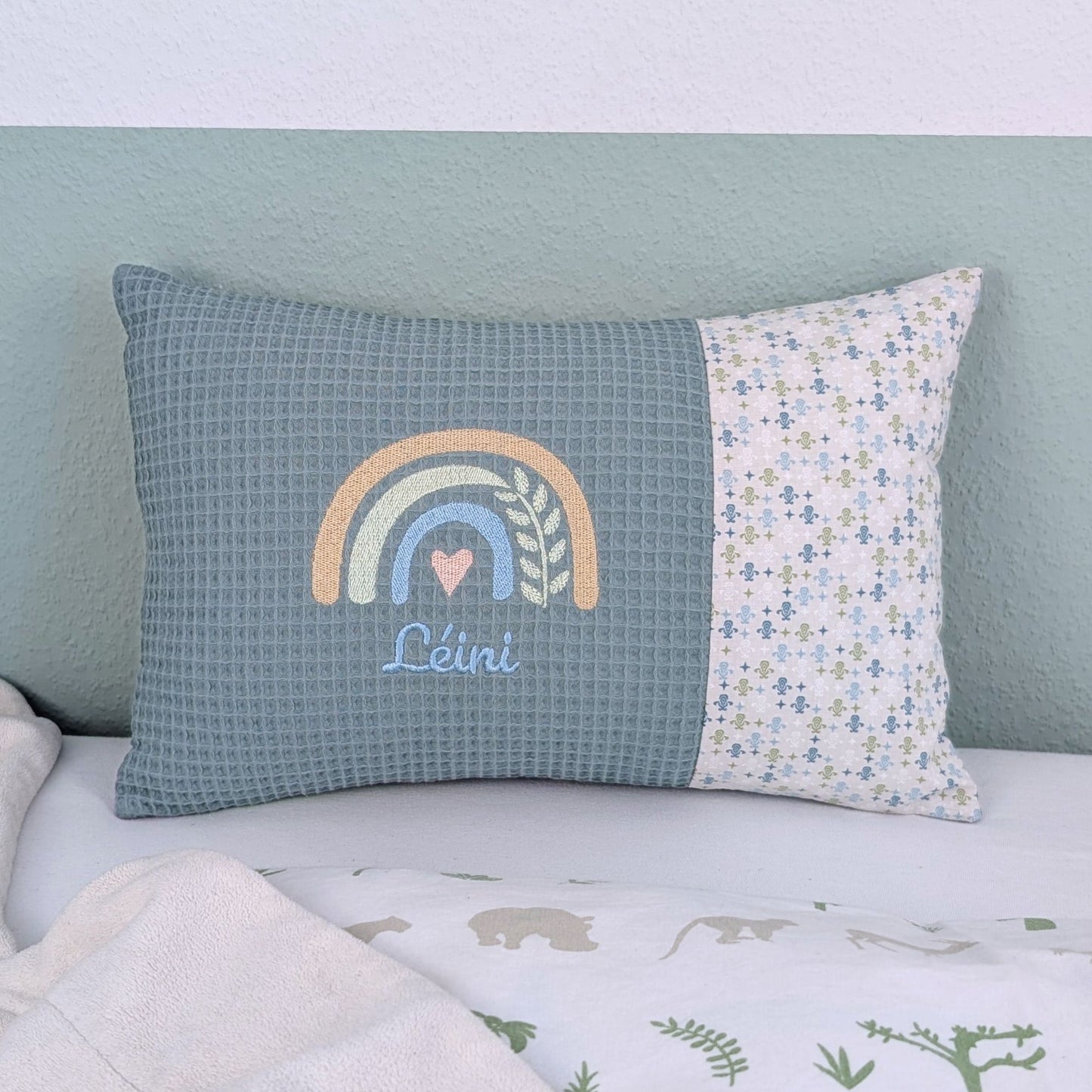 Personalisiertes Kissen mit Name. Das Kissen hat eine grüne Waffelstoff-Seite mit einer Stickerei eines Regenbogens in Gold, Grün und Blau, ergänzt durch ein Herz- und Ährenmotiv. Darunter ist der Name in blauer Schrift gestickt. Die rechte Seite des Kissens besteht aus einem gemusterten Stoff.