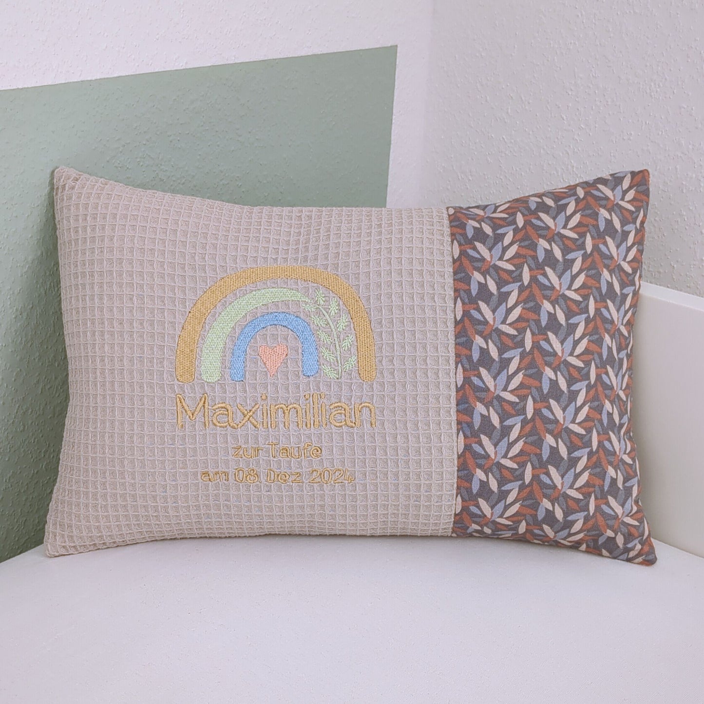 Personalisiertes Taufkissen mit Namen. Die linke Seite des Kissens besteht aus cremefarbenem Waffelstoff mit einer Stickerei eines Regenbogens in Gelb, Grün und Blau sowie einem kleinen Herzmotiv. Darunter steht in goldener Schrift 'zur Taufe am 08. Dez. 2024'. Die rechte Seite ist aus einem gemusterten Stoff mit Blättern in Erdtönen. Das Kissen liegt auf einer weißen Oberfläche und lehnt an eine grün-weiße Wand."