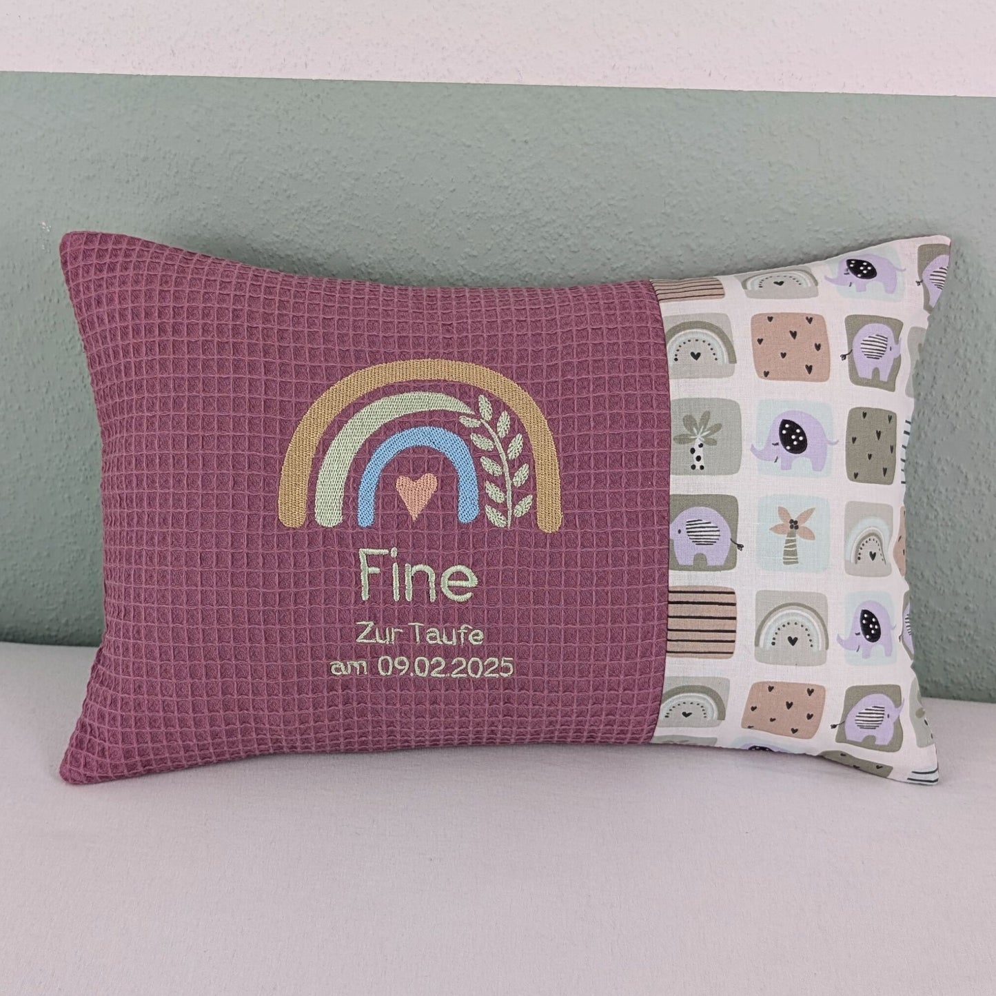 Dekoratives Taufkissen mit gesticktem Namen 'Fine'. Das Kissen hat eine lavendelfarbene Waffelstoff-Seite mit einer Stickerei eines Regenbogens in Beige, Blau und Gold, ergänzt durch ein Herz- und Ährenmotiv. Darunter steht 'Zur Taufe am 09.02.2025' in grüner Schrift. Die rechte Seite besteht aus einem gemusterten Stoff mit Elefanten, Regenbögen und abstrakten Formen in Pastellfarben. Das Kissen liegt auf einer hellen Oberfläche, an eine grün-weiße Wand gelehnt."
