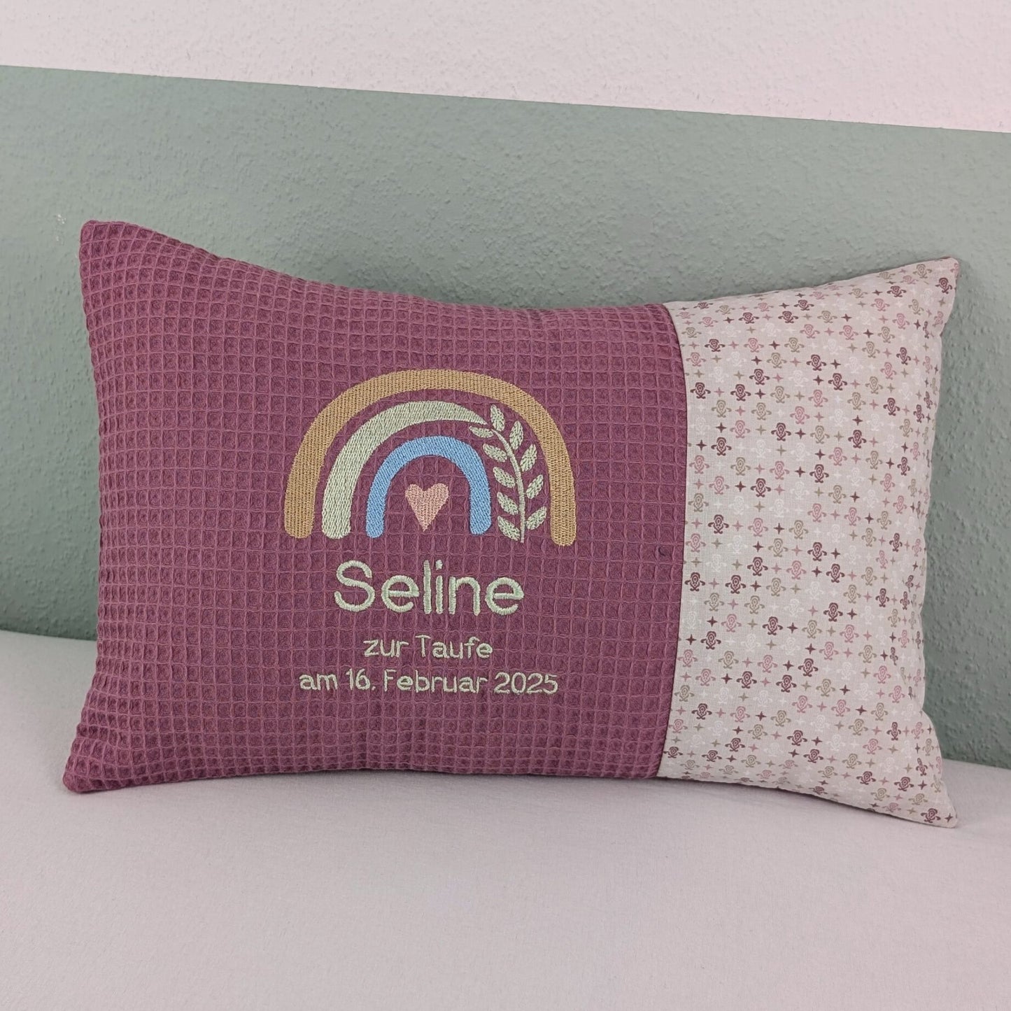 Dekoratives Taufkissen mit gesticktem Namen. Das Kissen hat eine lavendelfarbene Waffelstoff-Seite mit einer Stickerei eines Regenbogens in Beige, Blau und Gold, ergänzt durch ein Herz- und Ährenmotiv. Darunter steht 'Zur Taufe am 16.Februar 2025' in grüner Schrift. Die rechte Seite besteht aus einem gemusterten Stoff. Das Kissen liegt auf einer hellen Oberfläche, an eine grün-weiße Wand gelehnt."