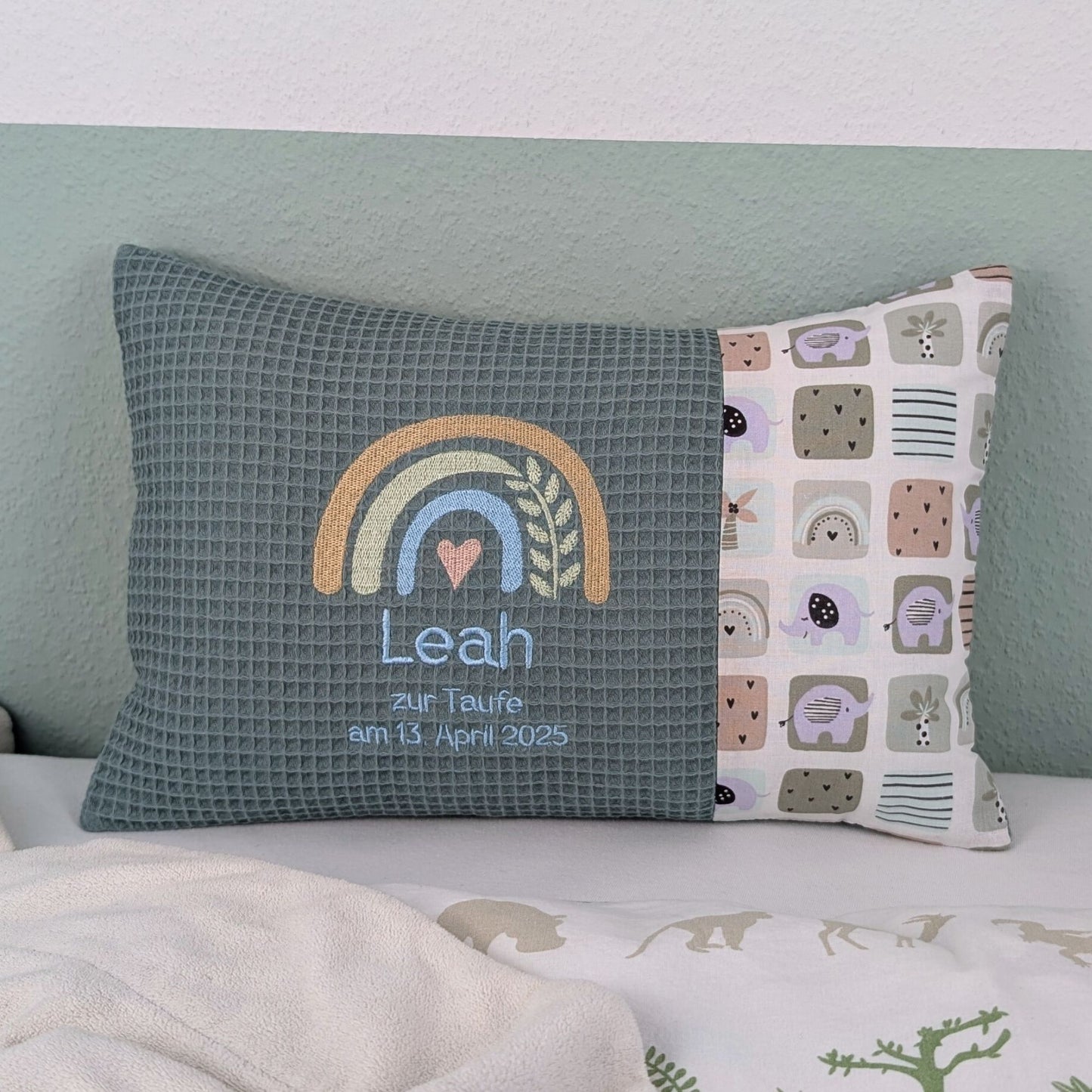 Dekoratives Taufkissen mit gesticktem Namen. Das Kissen hat eine türkisen Waffelstoff-Seite mit einer Stickerei eines Regenbogens in Beige, Blau und Gold, ergänzt durch ein Herz- und Ährenmotiv. Darunter steht, der Name vom Kind, 'Zur Taufe am 13.April 2025' in grüner Schrift. Die rechte Seite besteht aus einem gemusterten Stoff. Das Kissen liegt auf einer hellen Oberfläche, an eine grün-weiße Wand gelehnt."
