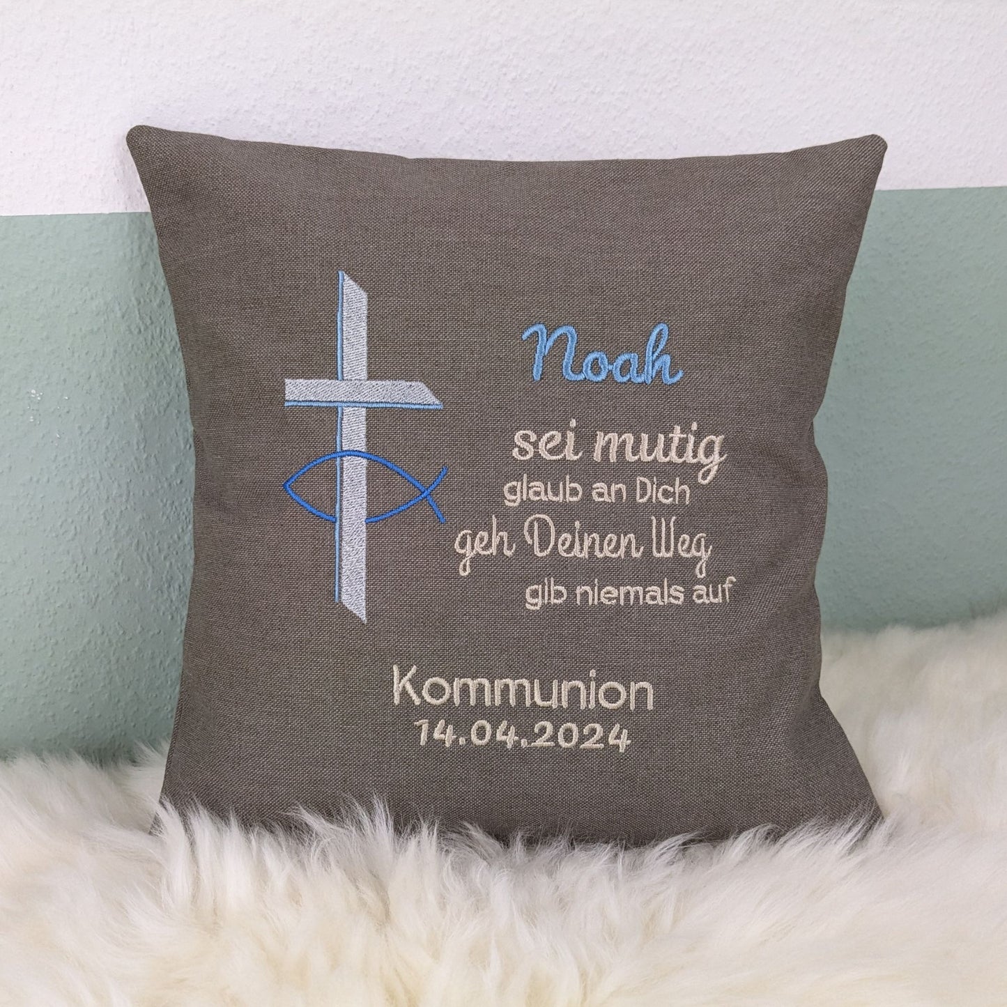 "Besticktes Kissen zur Kommunion. Das Kissen ist grau und zeigt eine Stickerei mit einem christlichen Kreuz und einem Fischsymbol in Blau und Silber. Der Name 'Noah' ist in blauer Schrift gestickt, gefolgt von einem ermutigenden Spruch in Beige: 'Sei mutig, glaub an Dich, geh Deinen Weg, gib niemals auf'. Darunter steht 'Kommunion 14.04.2024' in weißer Schrift. Das Kissen liegt auf einem weißen, flauschigen Untergrund."