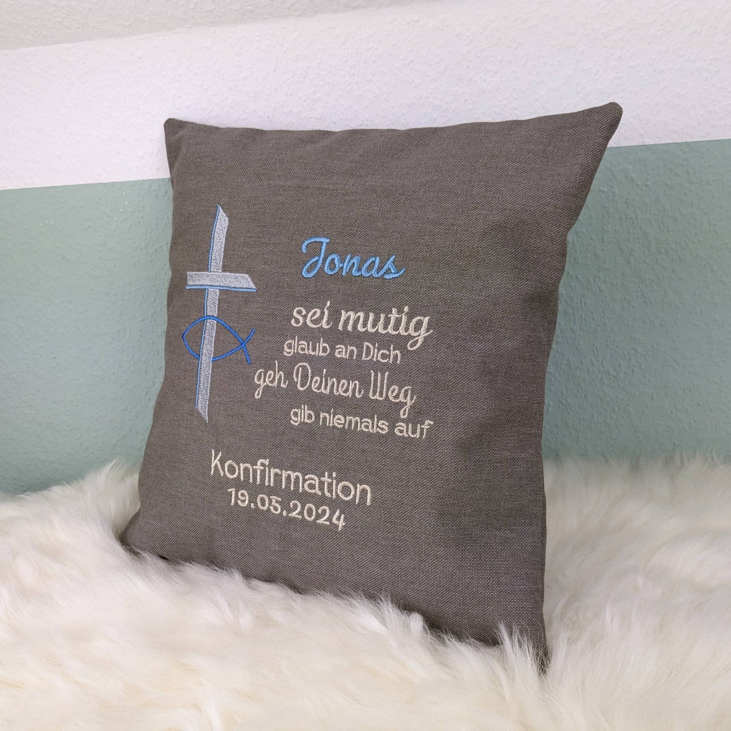 "Besticktes Kissen zur Konfirmation. Das Kissen ist grau und zeigt eine Stickerei mit einem christlichen Kreuz und einem Fischsymbol in blau und Silber. Der Name 'Jonas' ist in blauer Schrift gestickt, gefolgt von einem ermutigenden Spruch in Beige: 'Sei mutig, glaub an Dich, geh Deinen Weg, gib niemals auf'. Darunter steht 'Konfirmation 19.05.2024' in weißer Schrift. Das Kissen liegt auf einem weißen, flauschigen Untergrund."