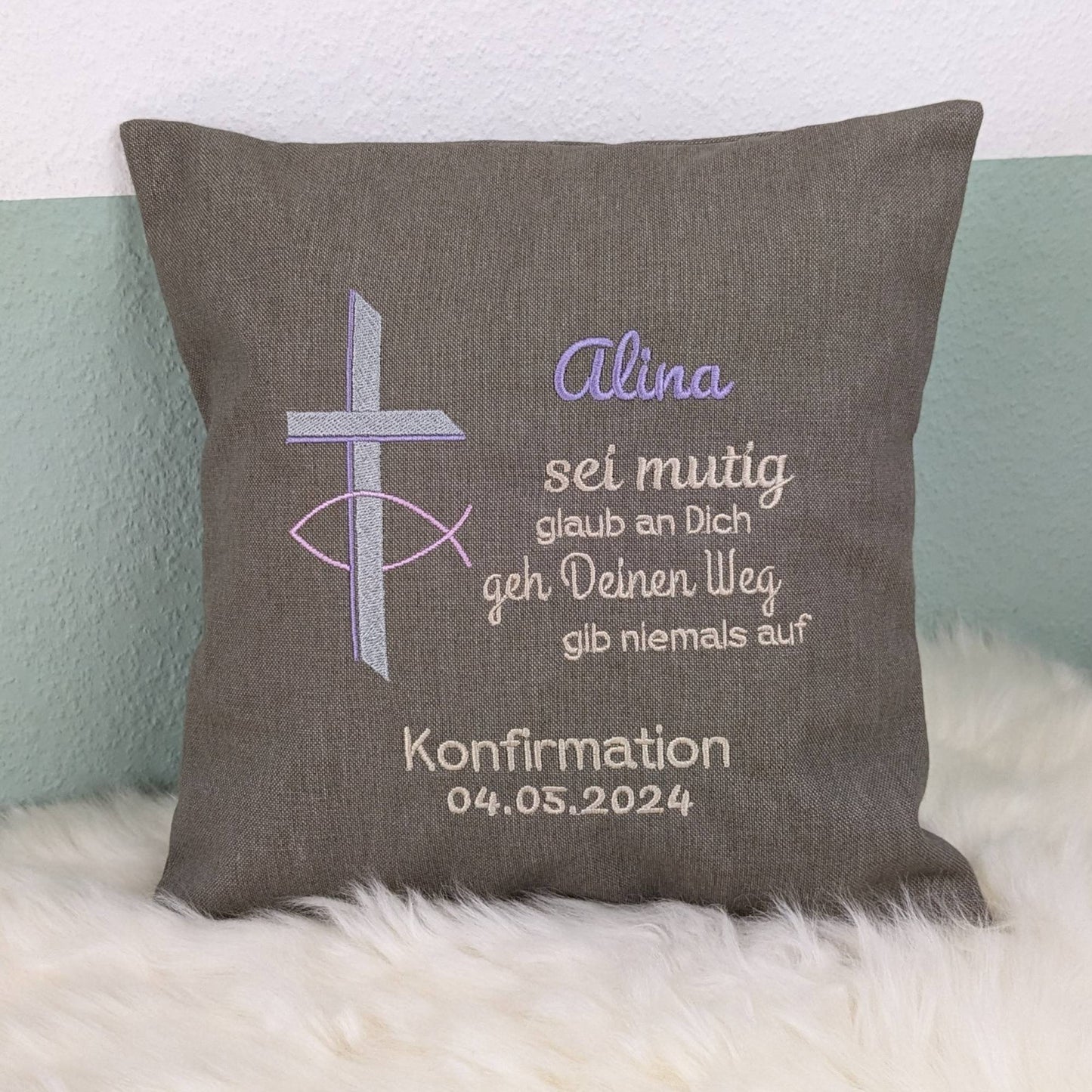"Besticktes Kissen zur Konfirmation. Das Kissen ist grau und zeigt eine Stickerei mit einem christlichen Kreuz und einem Fischsymbol in rosa, lavendel und Silber. Der Name 'Alina' ist in lila Schrift gestickt, gefolgt von einem ermutigenden Spruch in Beige: 'Sei mutig, glaub an Dich, geh Deinen Weg, gib niemals auf'. Darunter steht 'Konfirmation 04.05.2024' in weißer Schrift. Das Kissen liegt auf einem weißen, flauschigen Untergrund."
