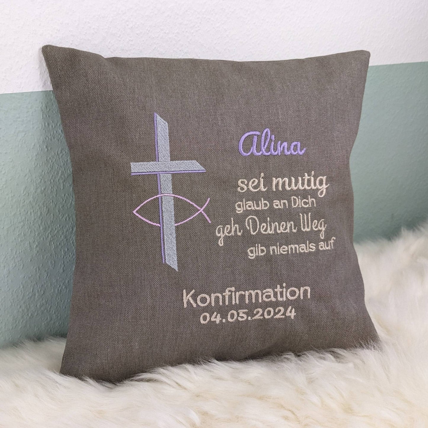 "Besticktes Kissen zur Konfirmation. Das Kissen ist grau und zeigt eine Stickerei mit einem christlichen Kreuz und einem Fischsymbol in rosa, lavendel und Silber. Der Name 'Alina' ist in lila Schrift gestickt, gefolgt von einem ermutigenden Spruch in Beige: 'Sei mutig, glaub an Dich, geh Deinen Weg, gib niemals auf'. Darunter steht 'Konfirmation 04.05.2024' in weißer Schrift. Das Kissen liegt auf einem weißen, flauschigen Untergrund."