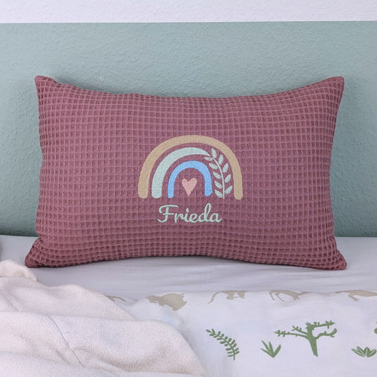 Kissen in der Größe 25 x 40 cm aus einem Waffelstoff in der Farbe altsosa. Mittig bestickt mit einem Regenbogen. Darunter steht ein Name.