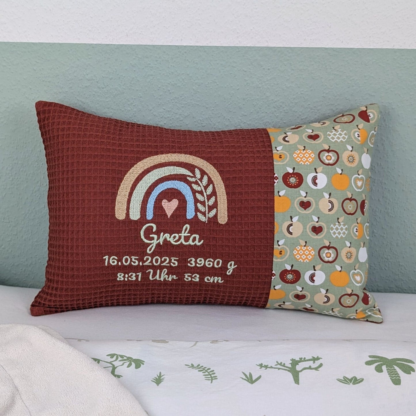 Personalisiertes Kissen mit Name und Geburtsdaten. Das Kissen hat eine rost Waffelstoff-Seite mit einer Stickerei eines Regenbogens in Gold, Grün und Blau, ergänzt durch ein Herz- und Ährenmotiv. Darunter ist der Name und die Geburtsdaten in grüner Schrift gestickt. Die rechte Seite des Kissens besteht aus einem gemusterten Stoff.