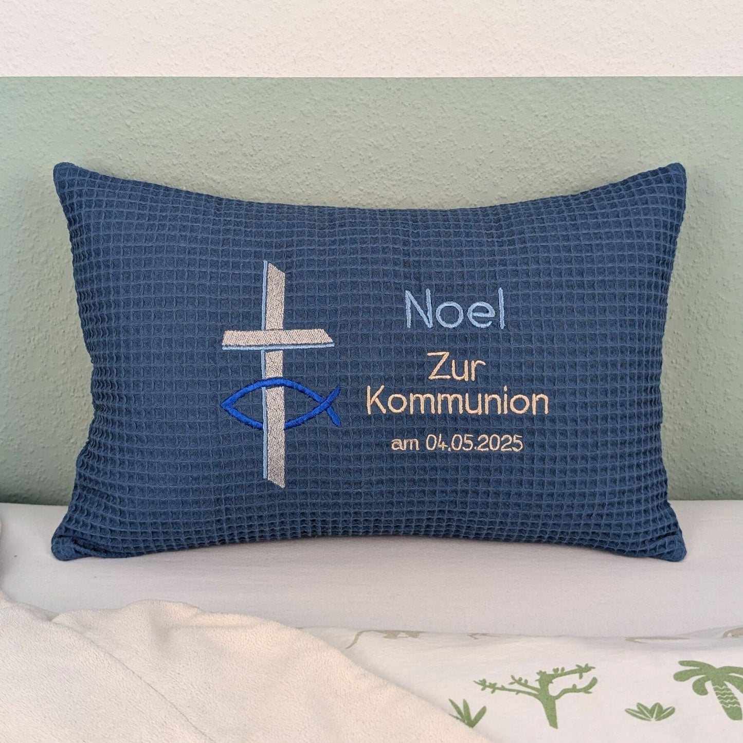 Rechteckiges Kissen in blauer Farbe mit Waffelstruktur liegt auf einem Bett. Auf dem Kissen sind ein gesticktes Kreuz und ein christliches Ichthys-Symbol (Fisch) sowie der Text „Noel – Zur Kommunion – am 04 .05.2025“ in verschiedenen Farben eingestickt. Das Kissen dient als personalisiertes Geschenk zur Erstkommunion.
