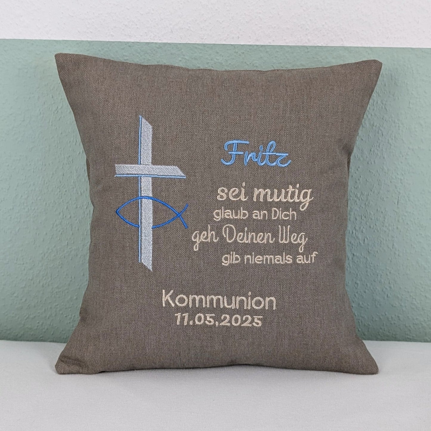"Besticktes Kissen zur Kommunion. Das Kissen ist grau und zeigt eine Stickerei mit einem christlichen Kreuz und einem Fischsymbol in Blau und Silber. Der Name 'Fritz' ist in blauer Schrift gestickt, gefolgt von einem ermutigenden Spruch in Beige: 'Sei mutig, glaub an Dich, geh Deinen Weg, gib niemals auf'. Darunter steht 'Kommunion 11.05.2025' in weißer Schrift. Das Kissen liegt auf einem weißen, Untergrund."