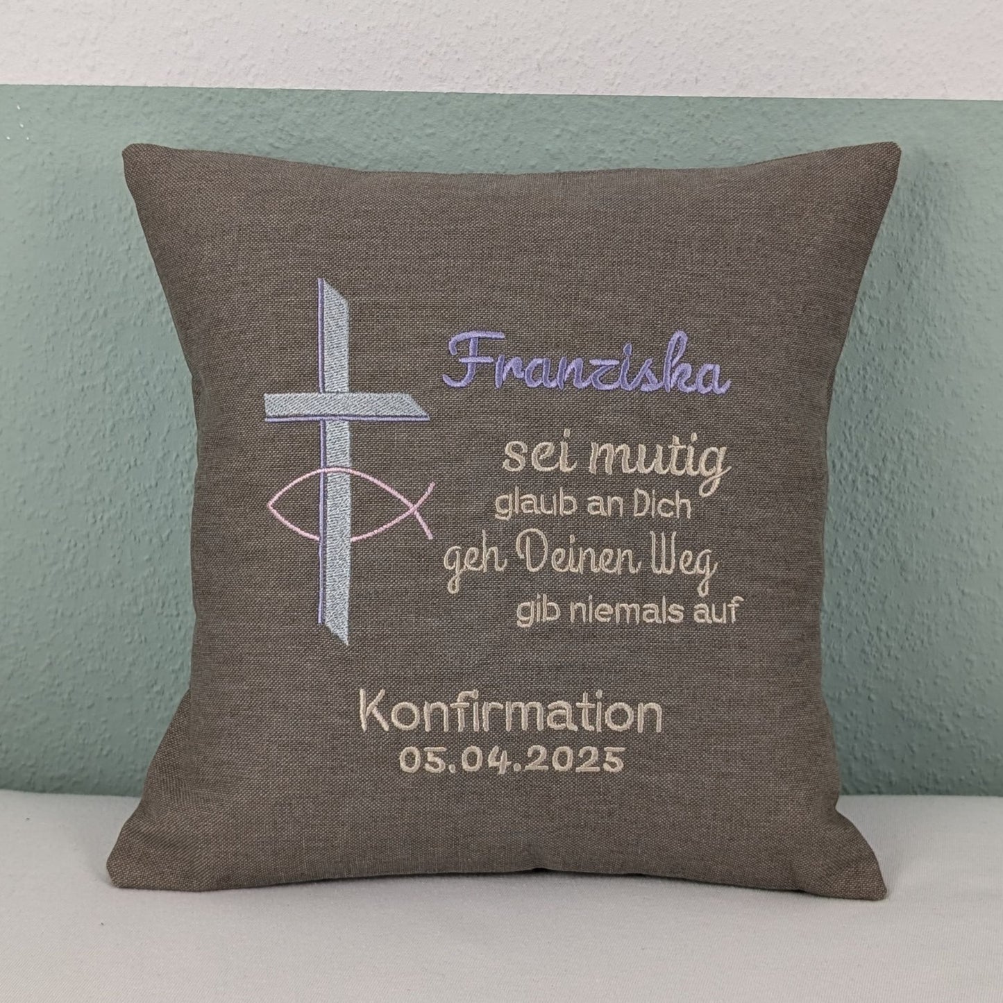 "Besticktes Kissen zur Konfirmation. Das Kissen ist grau und zeigt eine Stickerei mit einem christlichen Kreuz und einem Fischsymbol in lavendel und Silber. Der Name 'Noah' ist in blauer Schrift gestickt, gefolgt von einem ermutigenden Spruch in Beige: 'Sei mutig, glaub an Dich, geh Deinen Weg, gib niemals auf'. Darunter steht 'Konfirmation 05.04.2025' in weißer Schrift. Das Kissen liegt auf einem weißen, Untergrund."