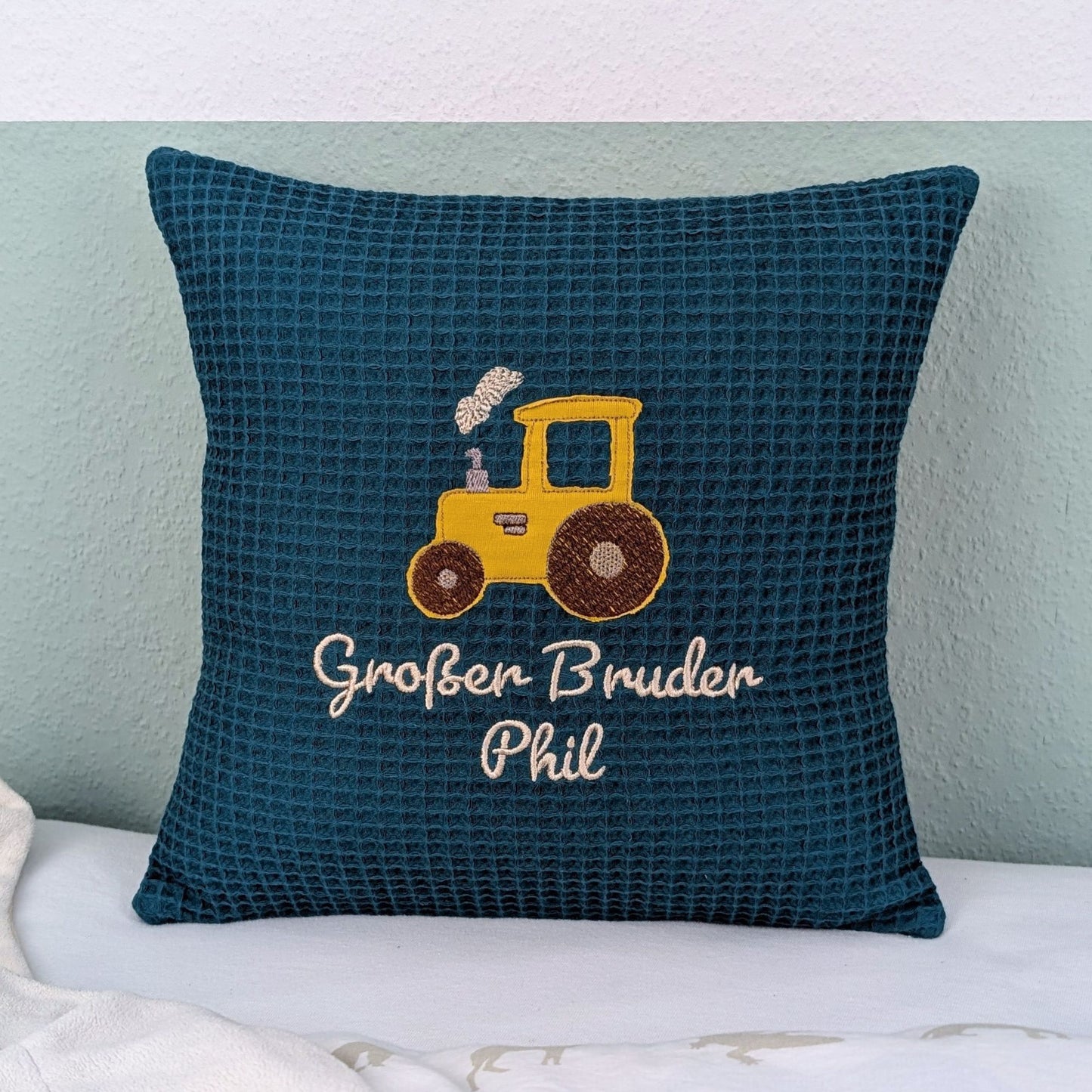 Ein Kissen in 30x30 cm aus Waffelstoff in der Farbe petrol. Bestickt mit einem Traktor in gelb mit Rauchendem Schornstein und darunter ist in weißer Schrift "Großer Bruder Phil" gestickt.