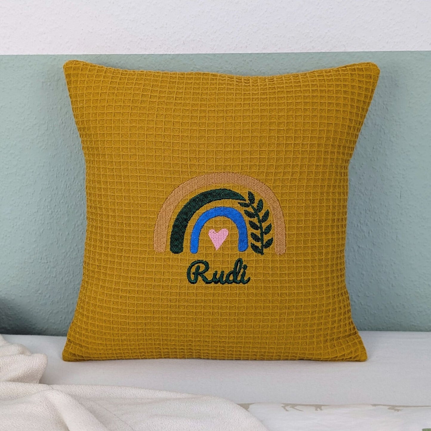 Kissen in der Größe 30 x 30 cm, aus Waffelstoff in der Farbe ocker, bestickt mit einem Regenbogen und darunter steht mittig eine Name.