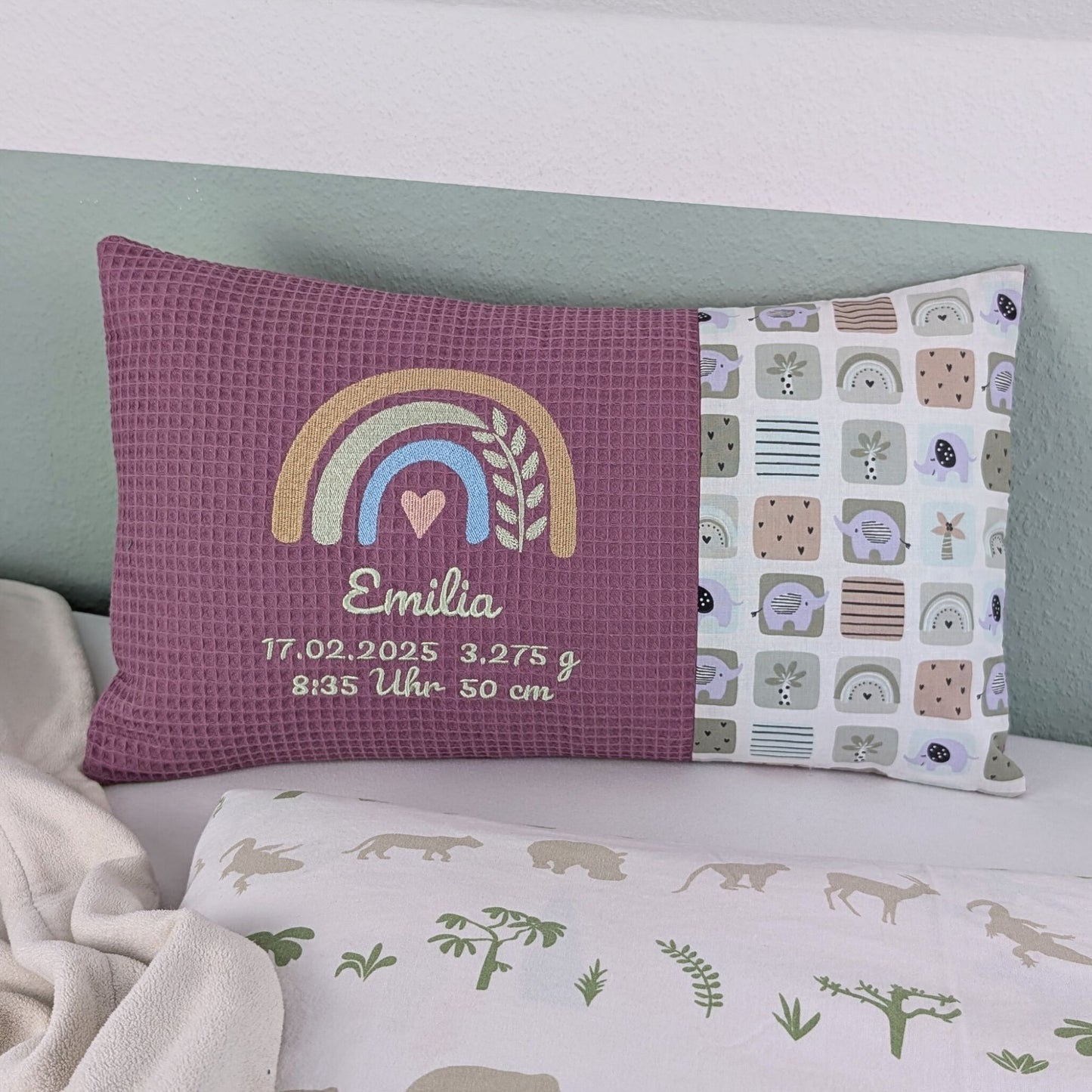 Dekoratives Geburtskissen mit gesticktem Namen und Geburtsdaten. Das Kissen hat eine alt rosa Waffelstoff-Seite mit einer Stickerei eines Regenbogens in Beige, Blau und Gold, ergänzt durch ein Herz- und Ährenmotiv. Darunter steht, der Name vom Kind, sowie Geburtsdatum Größe Gewicht und Uhrzeit in grüner Schrift. Die rechte Seite besteht aus einem gemusterten Stoff. Das Kissen liegt auf einer hellen Oberfläche, an eine grün-weiße Wand gelehnt."
