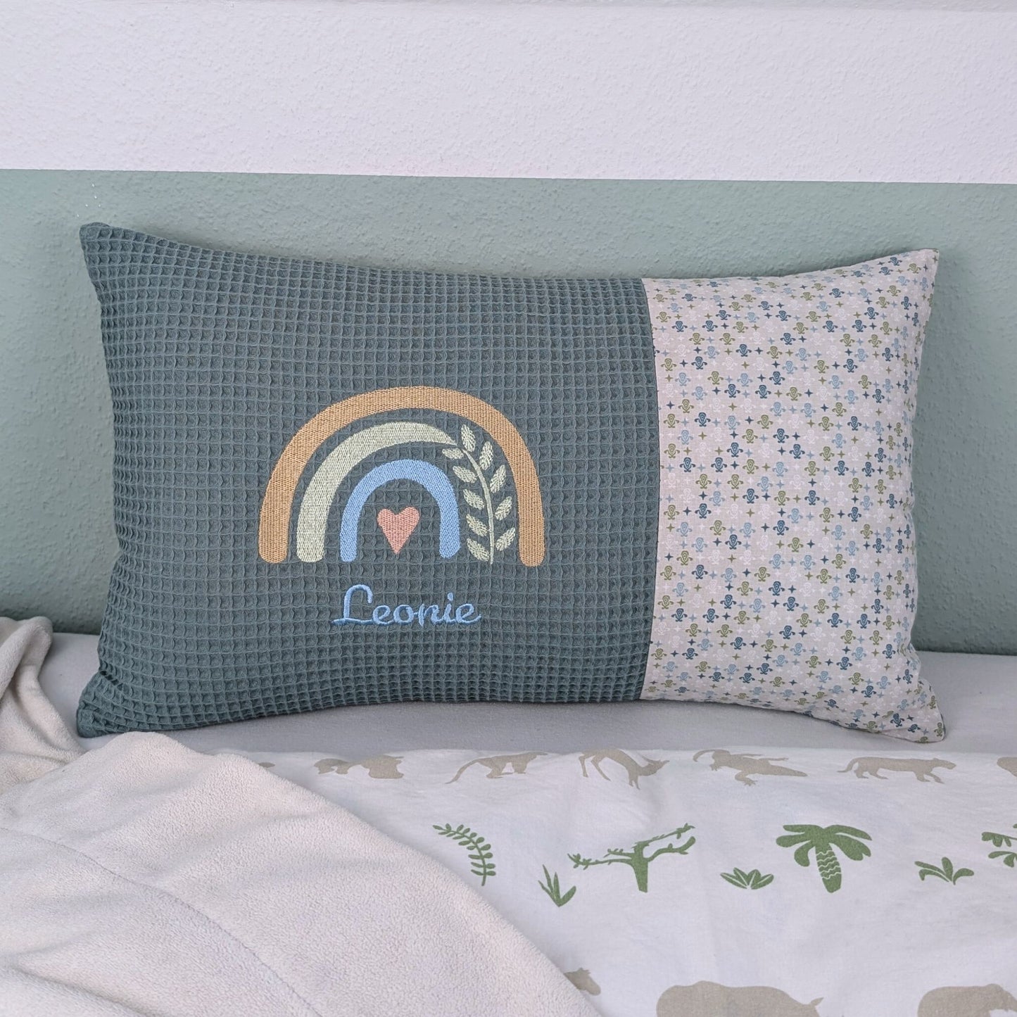 Dekoratives Namenskissen mit gesticktem Namen. Das Kissen hat eine türkisen Waffelstoff-Seite mit einer Stickerei eines Regenbogens in Beige, Blau und Gold, ergänzt durch ein Herz- und Ährenmotiv. Darunter steht, der Name vom Kind in grüner Schrift. Die rechte Seite besteht aus einem gemusterten Stoff. Das Kissen liegt auf einer hellen Oberfläche, an eine grün-weiße Wand gelehnt."