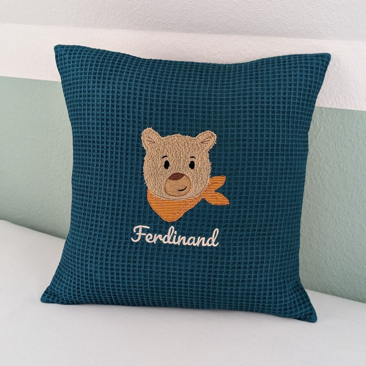 Personalisiertes Kissen in Petrol mit gesticktem Bärenmotiv. Der Bär hat eine beige, flauschige Struktur, schwarze Augen und eine braune Nase. Um seinen Hals trägt er ein orange-gestreiftes Halstuch. Darunter ist der Name in weißer Schrift gestickt. Das Kissen hat eine waffelartige Stoffstruktur und liegt auf einer weißen Oberfläche, an eine grün-weiße Wand gelehnt.