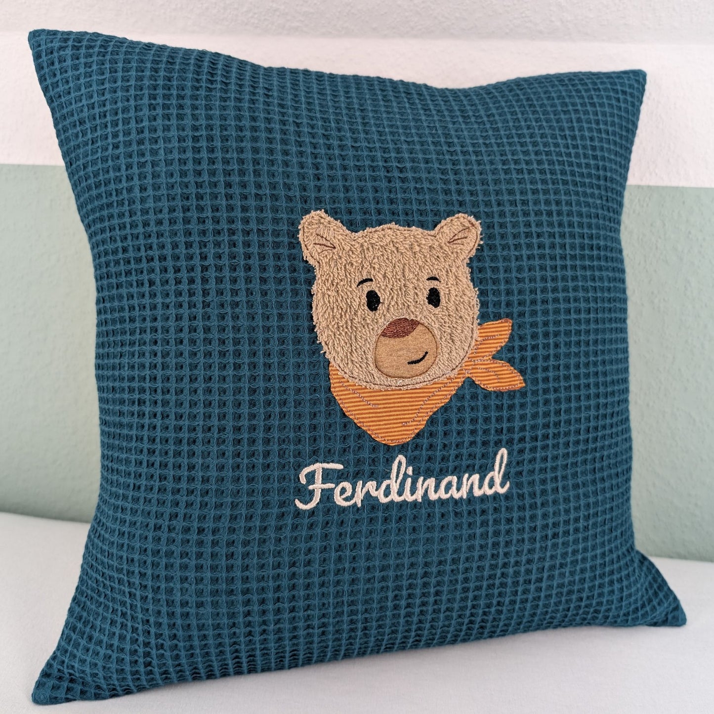 Personalisiertes Kissen in Petrol mit gesticktem Bärenmotiv. Der Bär hat eine beige, flauschige Struktur, schwarze Augen und eine braune Nase. Um seinen Hals trägt er ein orange-gestreiftes Halstuch. Darunter ist der Name in weißer Schrift gestickt. Das Kissen hat eine waffelartige Stoffstruktur und liegt auf einer weißen Oberfläche, an eine grün-weiße Wand gelehnt.