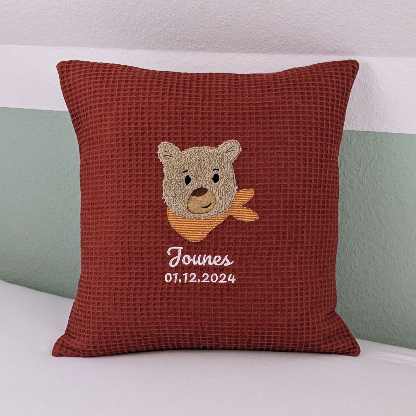 Kissen in der Größe 40x40 cm aus einem Waffelstoff in der Farbe rost. Es ist bestickt mit einem Bärenkopf der ein orangenes Halstuch um hat. Darunter ist der der Name Jounes und Geburtsdatum in weißer Schrift gestickt.