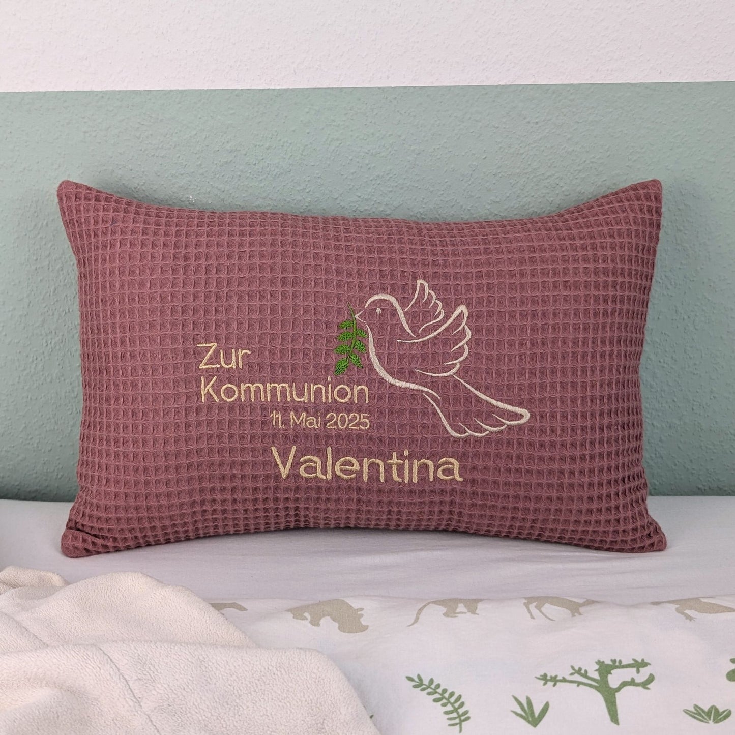 Rechteckiges, altrosa farbendes Kissen aus Waffelstoff liegt auf einem Bett. Auf dem Kissen ist eine weiße Taube mit einem grünen Olivenzweig im Schnabel sowie der gestickte Text „Zur Kommunion am 11. Mai 2025 – und der Name Valentina" zu sehen. Das Kissen dient als persönliches Geschenk zur Erstkommunion.