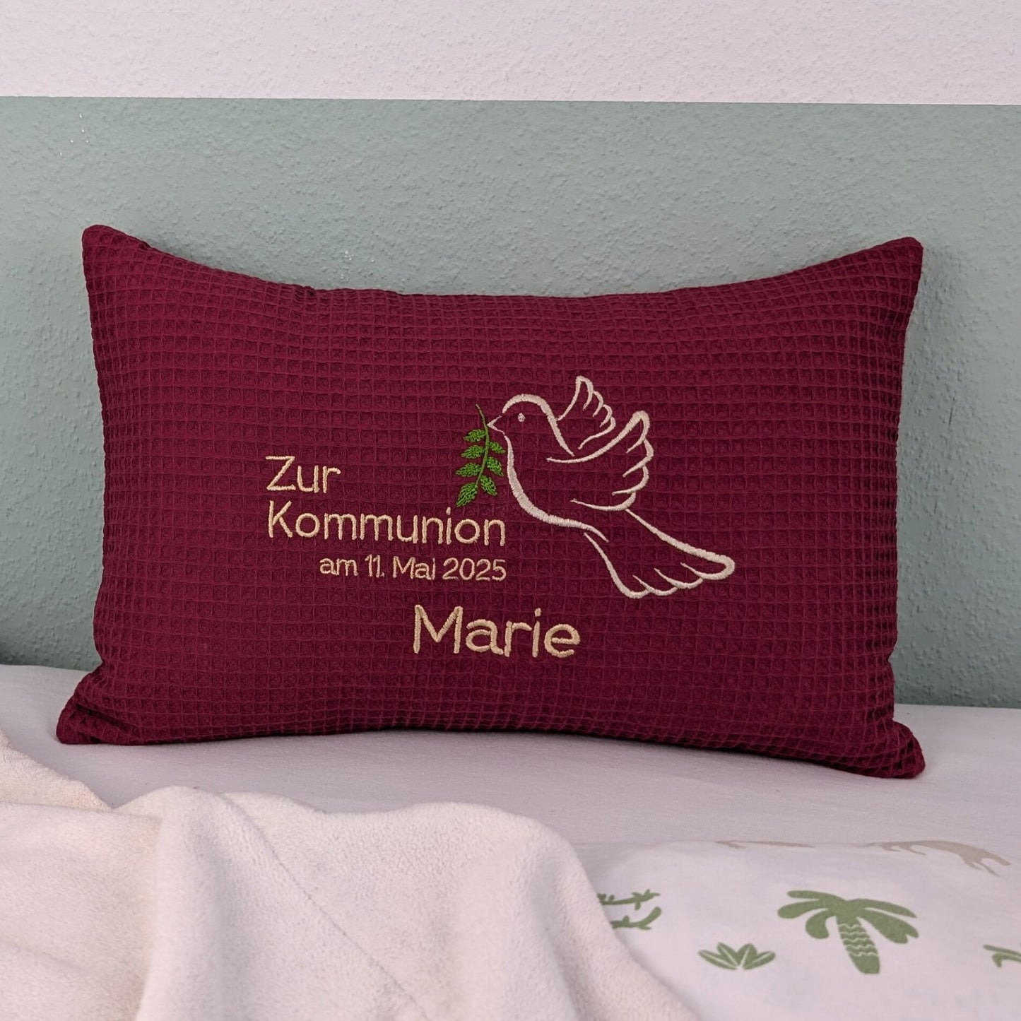 Rechteckiges, beerefarbendes Kissen aus Waffelstoff liegt auf einem Bett. Auf dem Kissen ist eine weiße Taube mit einem grünen Olivenzweig im Schnabel sowie der gestickte Text „Zur Kommunion am 11. Mai 2025 – und der Name Marie" zu sehen. Das Kissen dient als persönliches Geschenk zur Erstkommunion.