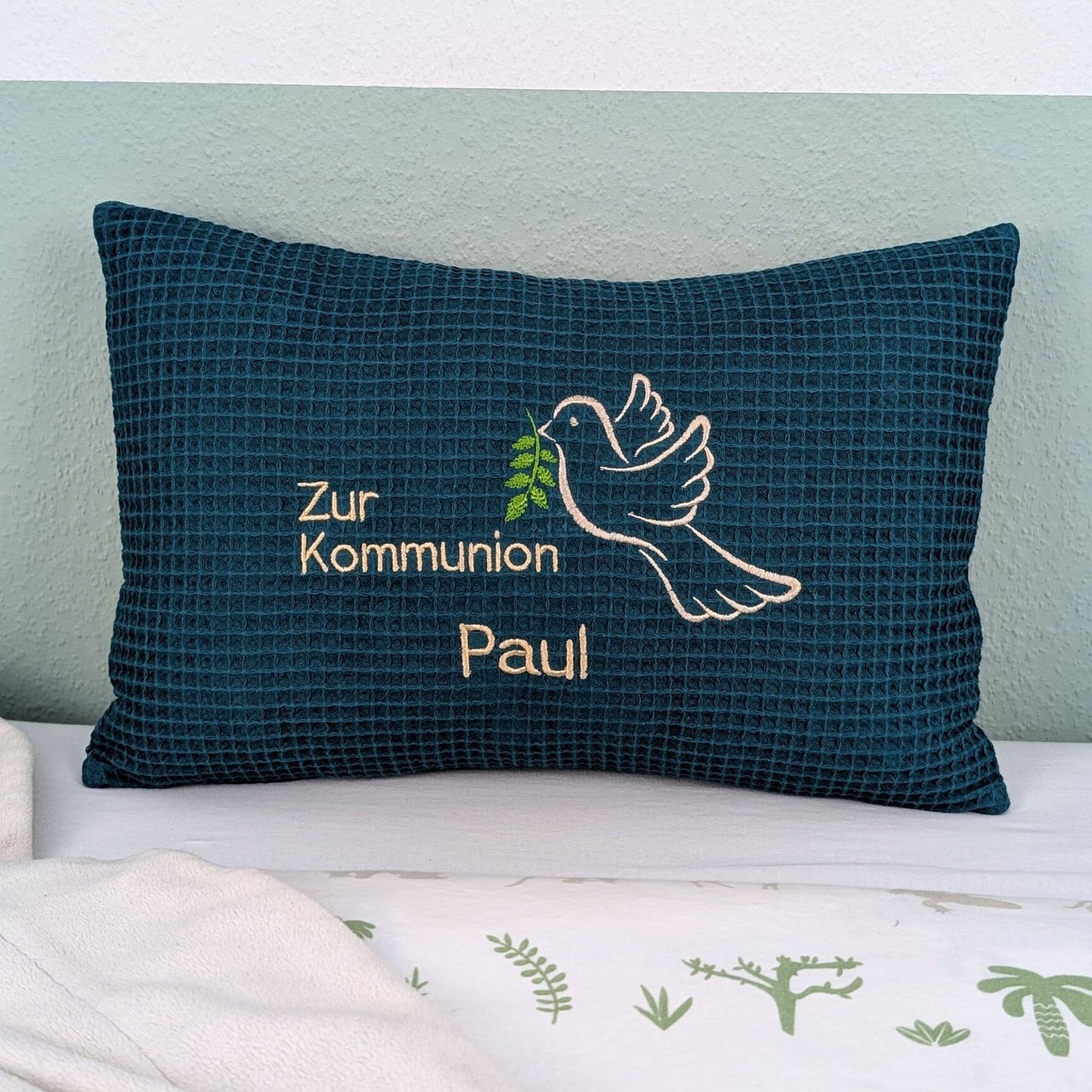 Rechteckiges, petrolfarbendes Kissen aus Waffelstoff liegt auf einem Bett. Auf dem Kissen ist eine weiße Taube mit einem grünen Olivenzweig im Schnabel sowie der gestickte Text „Zur Kommunion – und der Name Paul" zu sehen. Das Kissen dient als persönliches Geschenk zur Erstkommunion.