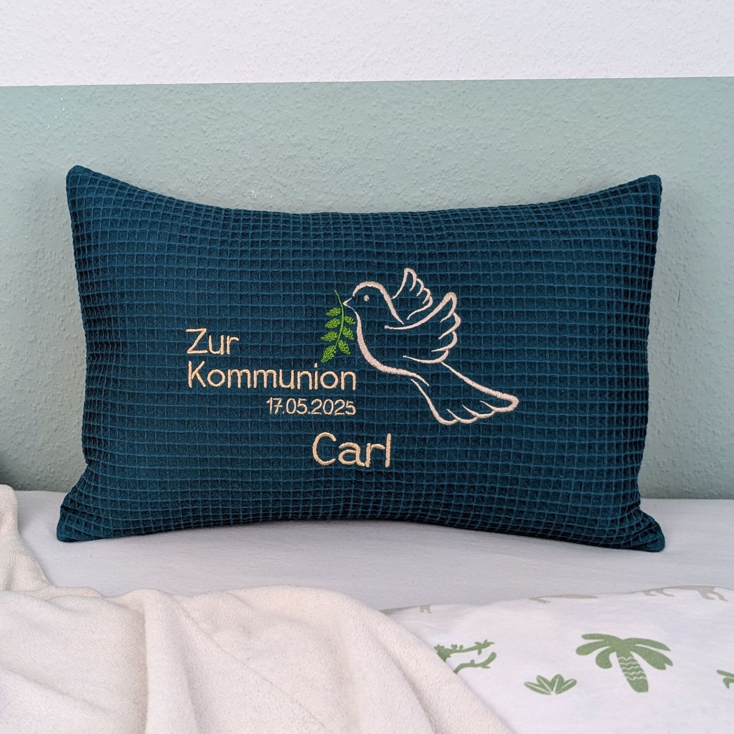 Rechteckiges, petrolfarbendes Kissen aus Waffelstoff liegt auf einem Bett. Auf dem Kissen ist eine weiße Taube mit einem grünen Olivenzweig im Schnabel sowie der gestickte Text „Zur Kommunion 17.05. 2025 – und der Name Carl" zu sehen. Das Kissen dient als persönliches Geschenk zur Erstkommunion.