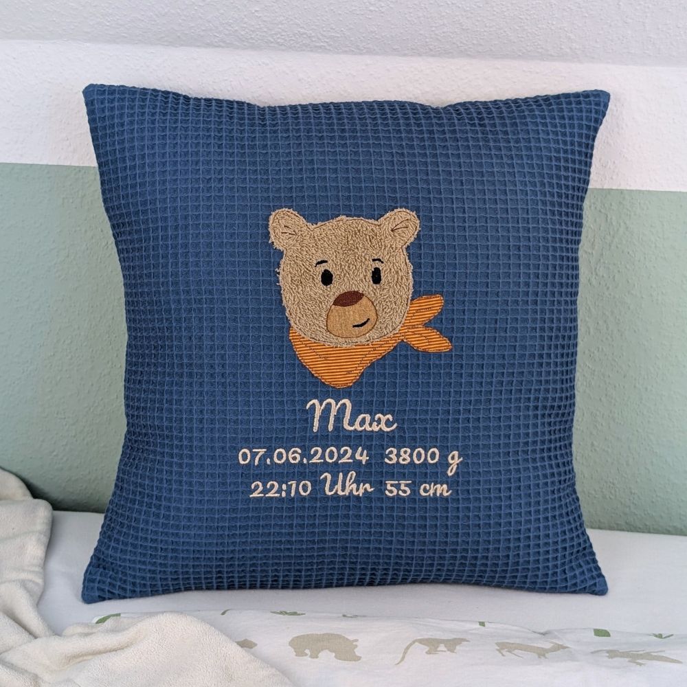 Kissen in der Größe 40x40 cm aus einem Waffelstoff in der Farbe Blau. Es ist bestickt mit einem Bärenkopf der ein orangenes Halstuch um hat. Darunter ist der der Name Max und Geburtsdaten wie Datum, Gewicht, Uhrzeit und Größe in weißer Schrift gestickt.