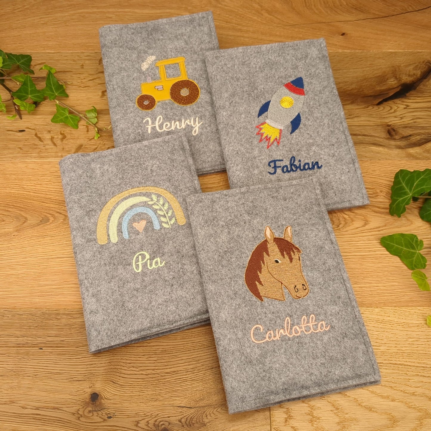 Vier Schutzhüllen für Untersuchungsheft aus grauem Filz, bestickt mit Rakete, Trecker, Pferd, Regenbogen und Name.