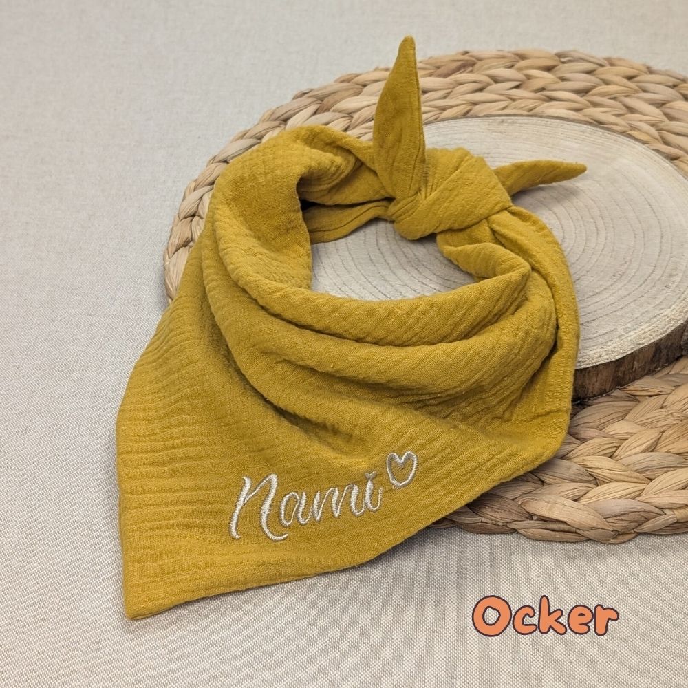 Es ist ein Halstuch aus Musselin bestickt mit dem Namen Nami und einem kleinen Herzen hinter dem Namen zu sehen. Das Tuch hat die Farbe senf.