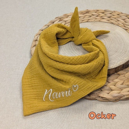 Es ist ein Halstuch aus Musselin bestickt mit dem Namen Nami und einem kleinen Herzen hinter dem Namen zu sehen. Das Tuch hat die Farbe senf.