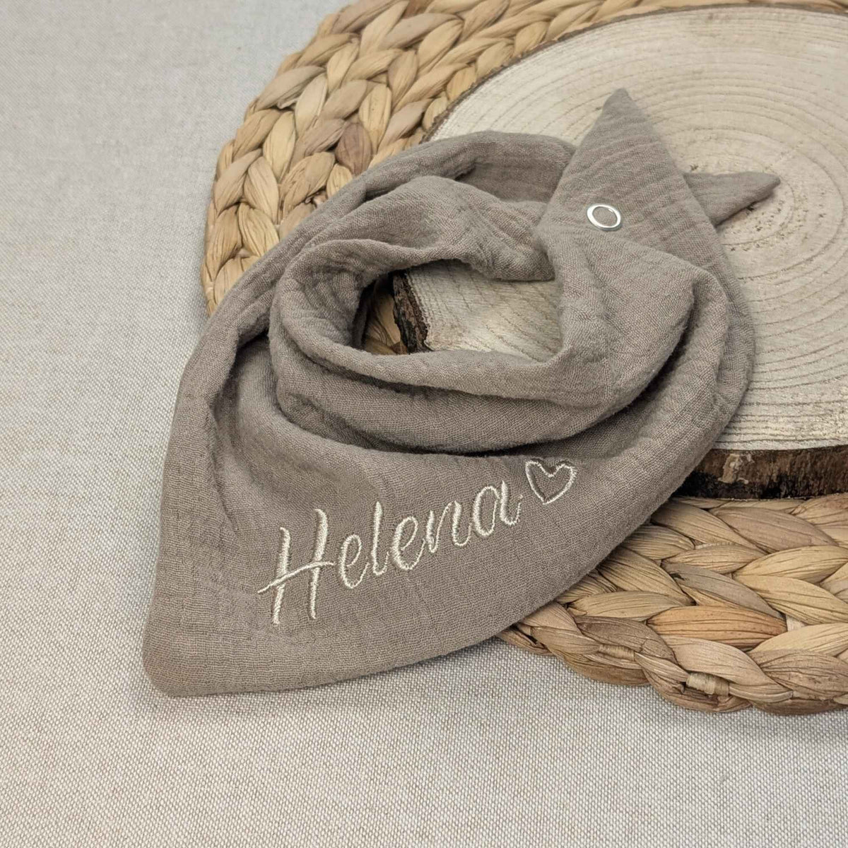 Es ist ein Halstuch aus Musselin bestickt mit dem Namen Helena und einem kleinen Herzen hinter dem Namen zu sehen. Das Tuch hat die Farbe taupe.