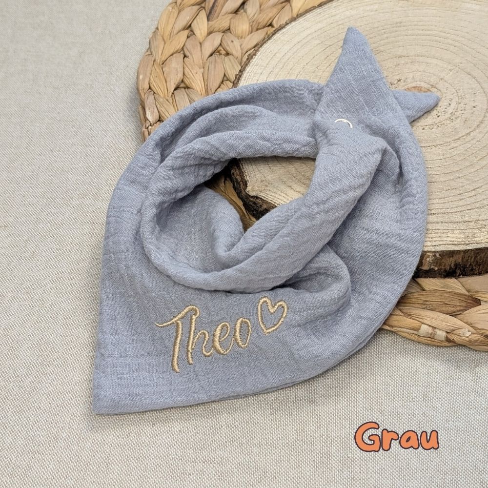 Es ist ein Halstuch aus Musselin bestickt mit dem Namen Nami und einem kleinen Herzen hinter dem Namen zu sehen. Das Tuch hat die Farbe grau.