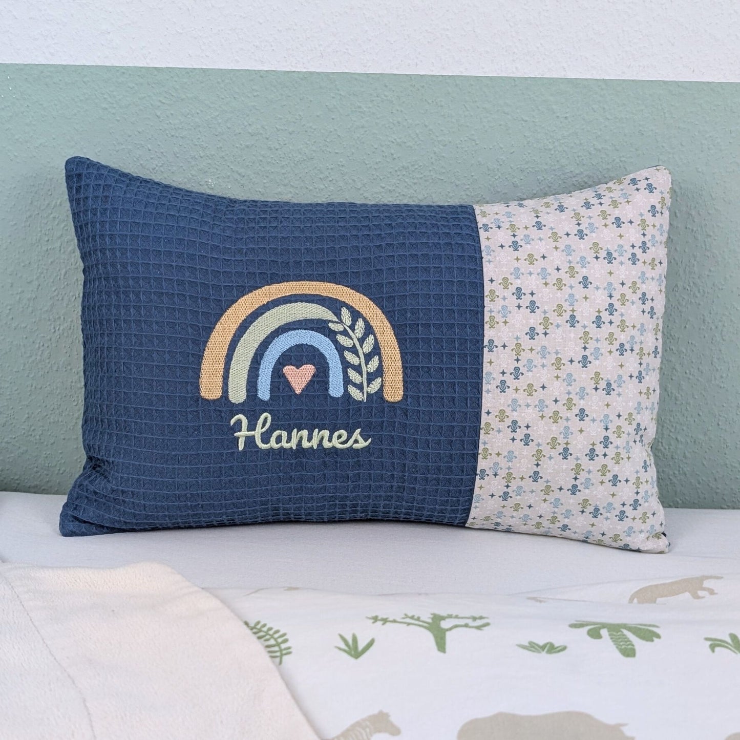Personalisiertes Kissen mit Name. Das Kissen hat eine blaue Waffelstoff-Seite mit einer Stickerei eines Regenbogens in Gold, Grün und Blau, ergänzt durch ein Herz- und Ährenmotiv. Darunter ist der Name in grüner Schrift gestickt. Die rechte Seite des Kissens besteht aus einem gemusterten Stoff.
