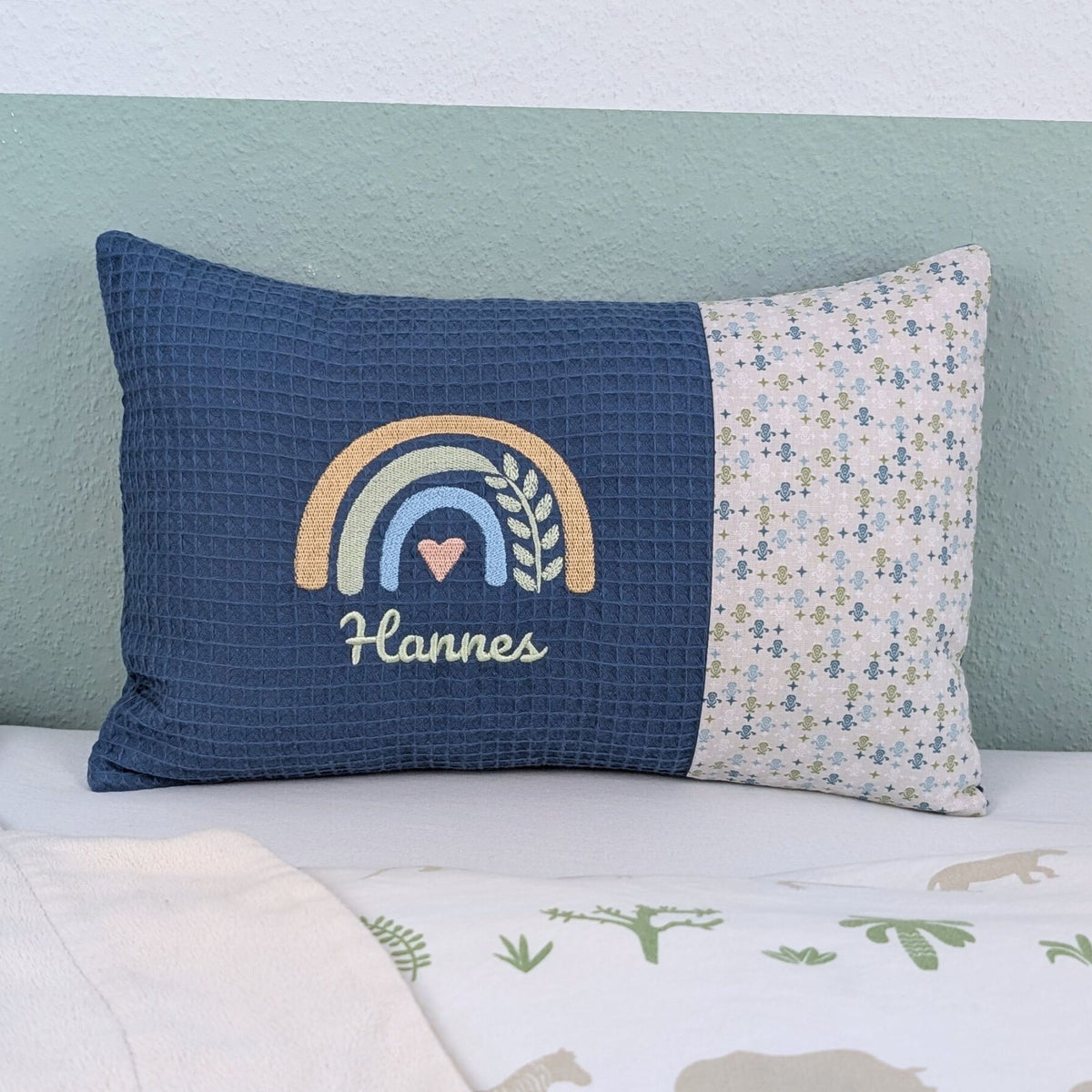 Personalisiertes Kissen mit Name. Das Kissen hat eine blaue Waffelstoff-Seite mit einer Stickerei eines Regenbogens in Gold, Grün und Blau, ergänzt durch ein Herz- und Ährenmotiv. Darunter ist der Name in grüner Schrift gestickt. Die rechte Seite des Kissens besteht aus einem gemusterten Stoff.