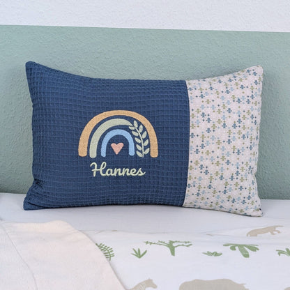 Personalisiertes Kissen mit Name. Das Kissen hat eine blaue Waffelstoff-Seite mit einer Stickerei eines Regenbogens in Gold, Grün und Blau, ergänzt durch ein Herz- und Ährenmotiv. Darunter ist der Name in grüner Schrift gestickt. Die rechte Seite des Kissens besteht aus einem gemusterten Stoff.