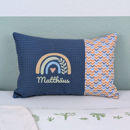 Personalisiertes Kissen mit Name. Das Kissen hat eine blaue Waffelstoff-Seite mit einer Stickerei eines Regenbogens in Gold, Grün und Blau, ergänzt durch ein Herz- und Ährenmotiv. Darunter ist der Name in grüner  Schrift gestickt. Die rechte Seite des Kissens besteht aus einem gemusterten Stoff.