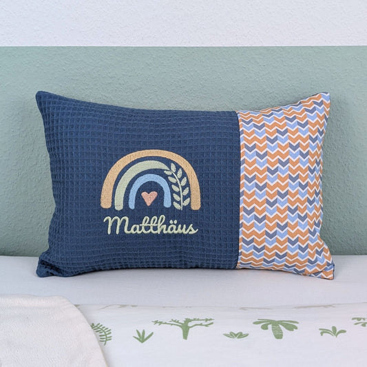 Personalisiertes Kissen mit Name. Das Kissen hat eine blaue Waffelstoff-Seite mit einer Stickerei eines Regenbogens in Gold, Grün und Blau, ergänzt durch ein Herz- und Ährenmotiv. Darunter ist der Name in grüner  Schrift gestickt. Die rechte Seite des Kissens besteht aus einem gemusterten Stoff.