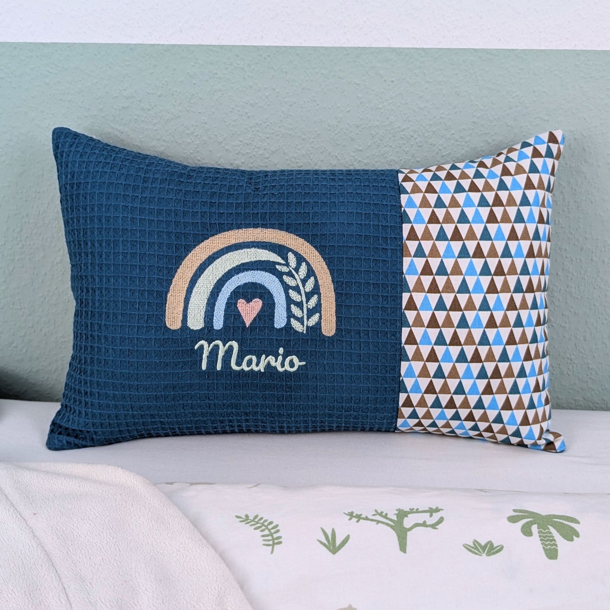 Personalisiertes Kissen mit Name. Das Kissen hat eine petrol Waffelstoff-Seite mit einer Stickerei eines Regenbogens in Gold, Grün und Blau, ergänzt durch ein Herz- und Ährenmotiv. Darunter ist der Name in grüner Schrift gestickt. Die rechte Seite des Kissens besteht aus einem gemusterten Stoff.