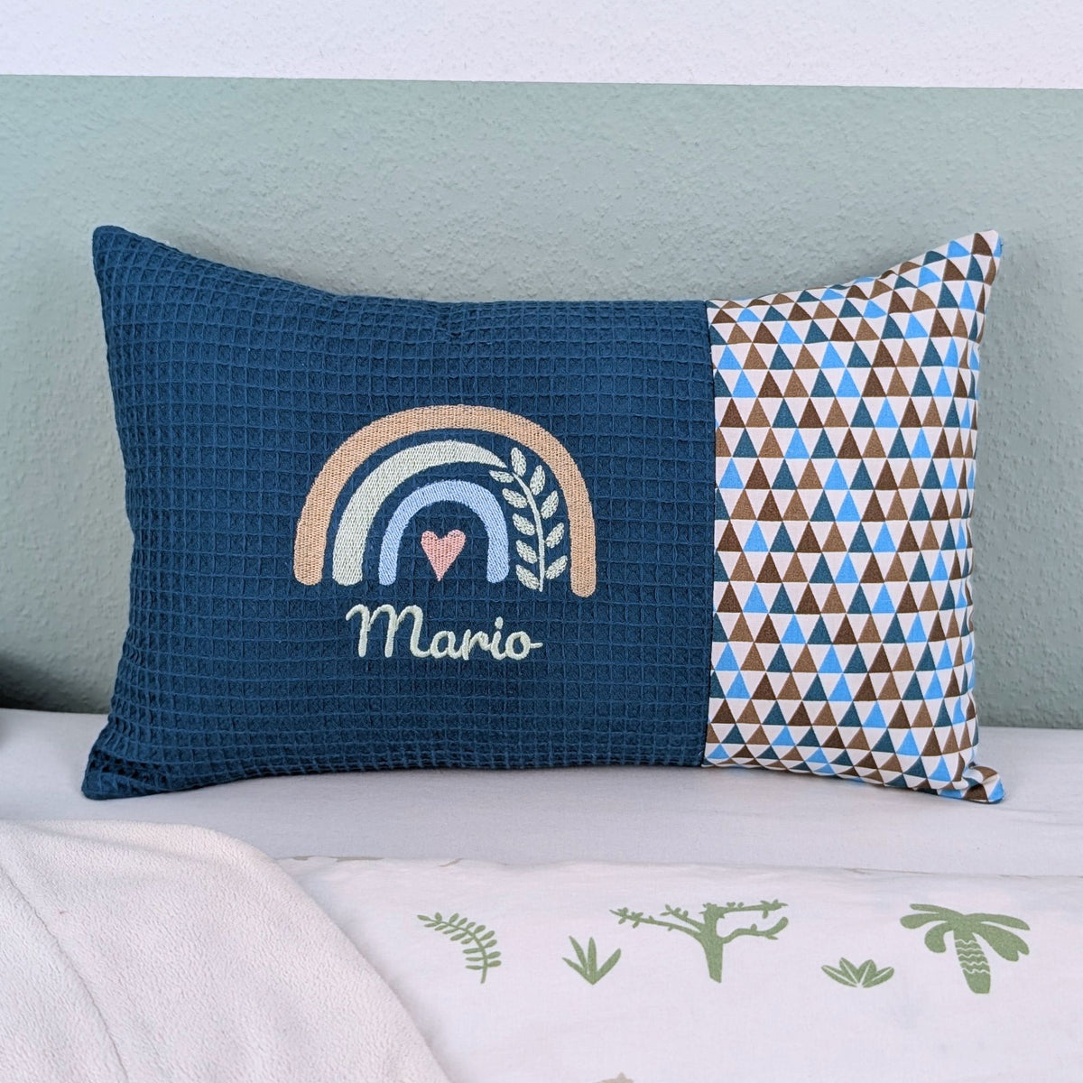 Personalisiertes Kissen mit Name. Das Kissen hat eine petrol Waffelstoff-Seite mit einer Stickerei eines Regenbogens in Gold, Grün und Blau, ergänzt durch ein Herz- und Ährenmotiv. Darunter ist der Name in grüner Schrift gestickt. Die rechte Seite des Kissens besteht aus einem gemusterten Stoff.