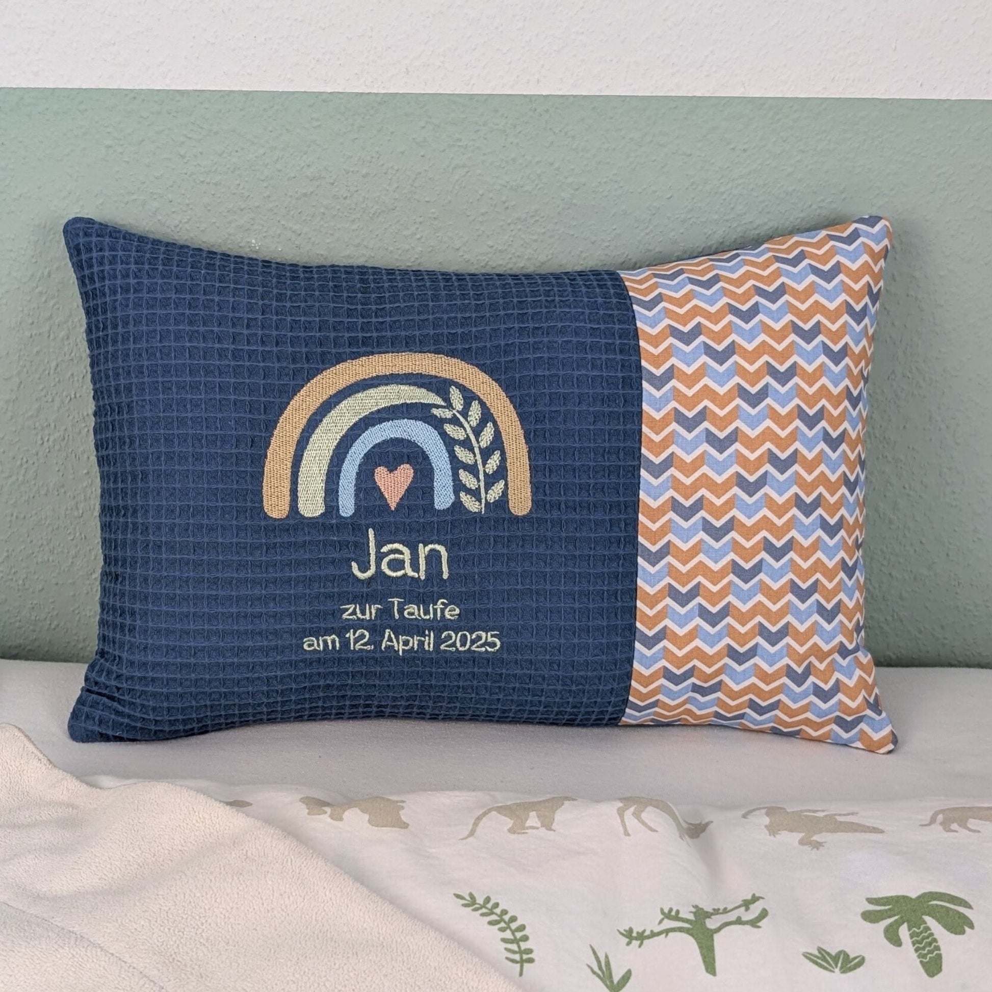 Dekoratives Taufkissen mit gesticktem Namen. Das Kissen hat eine blauen Waffelstoff-Seite mit einer Stickerei eines Regenbogens in Beige, Blau und Gold, ergänzt durch ein Herz- und Ährenmotiv. Darunter steht der Name vom Kind, 'Zur Taufe am 16.Februar 2025' in grüner Schrift. Die rechte Seite besteht aus einem gemusterten Stoff. Das Kissen liegt auf einer hellen Oberfläche, an eine grün-weiße Wand gelehnt."