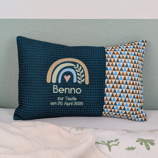 Dekoratives Taufkissen mit gesticktem Namen. Das Kissen hat eine petrolfarbenden Waffelstoff-Seite mit einer Stickerei eines Regenbogens in Beige, Blau und Gold, ergänzt durch ein Herz- und Ährenmotiv. Darunter steht der Name vom Kind, 'Zur Taufe am 20.April 2025' in mint grüner Schrift. Die rechte Seite besteht aus einem gemusterten Stoff. Das Kissen liegt auf einer hellen Oberfläche, an eine grün-weiße Wand gelehnt."