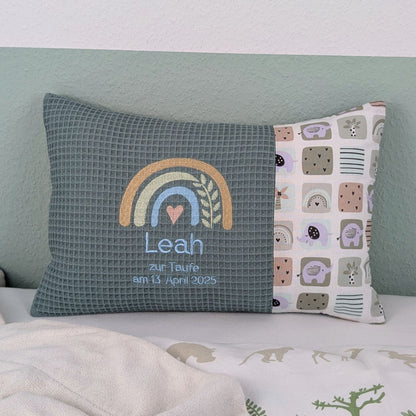 Dekoratives Taufkissen mit gesticktem Namen. Das Kissen hat eine türkisen Waffelstoff-Seite mit einer Stickerei eines Regenbogens in Beige, Blau und Gold, ergänzt durch ein Herz- und Ährenmotiv. Darunter steht, der Name vom Kind, 'Zur Taufe am 13.April 2025' in grüner Schrift. Die rechte Seite besteht aus einem gemusterten Stoff. Das Kissen liegt auf einer hellen Oberfläche, an eine grün-weiße Wand gelehnt."