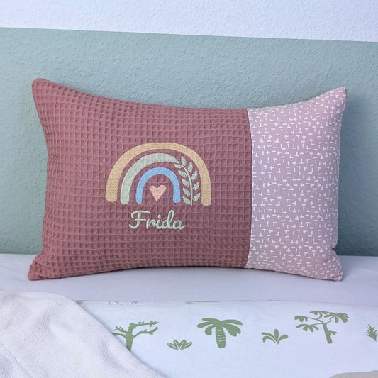 Personalisiertes Kissen mit Name. Das Kissen hat eine altrosa Waffelstoff-Seite mit einer Stickerei eines Regenbogens in Gold, Grün und Blau, ergänzt durch ein Herz- und Ährenmotiv. Darunter ist der Name in grüner Schrift gestickt. Die rechte Seite des Kissens besteht aus einem gemusterten Stoff.
