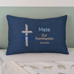 Rechteckiges Kissen in blauer Farbe mit Waffelstruktur liegt auf einem Bett. Auf dem Kissen sind ein gesticktes Kreuz und ein christliches Ichthys-Symbol (Fisch) sowie der Text „Mats – Zur Kommunion – 27.04.2025“ in verschiedenen Farben eingestickt. Das Kissen dient als personalisiertes Geschenk zur Erstkommunion.