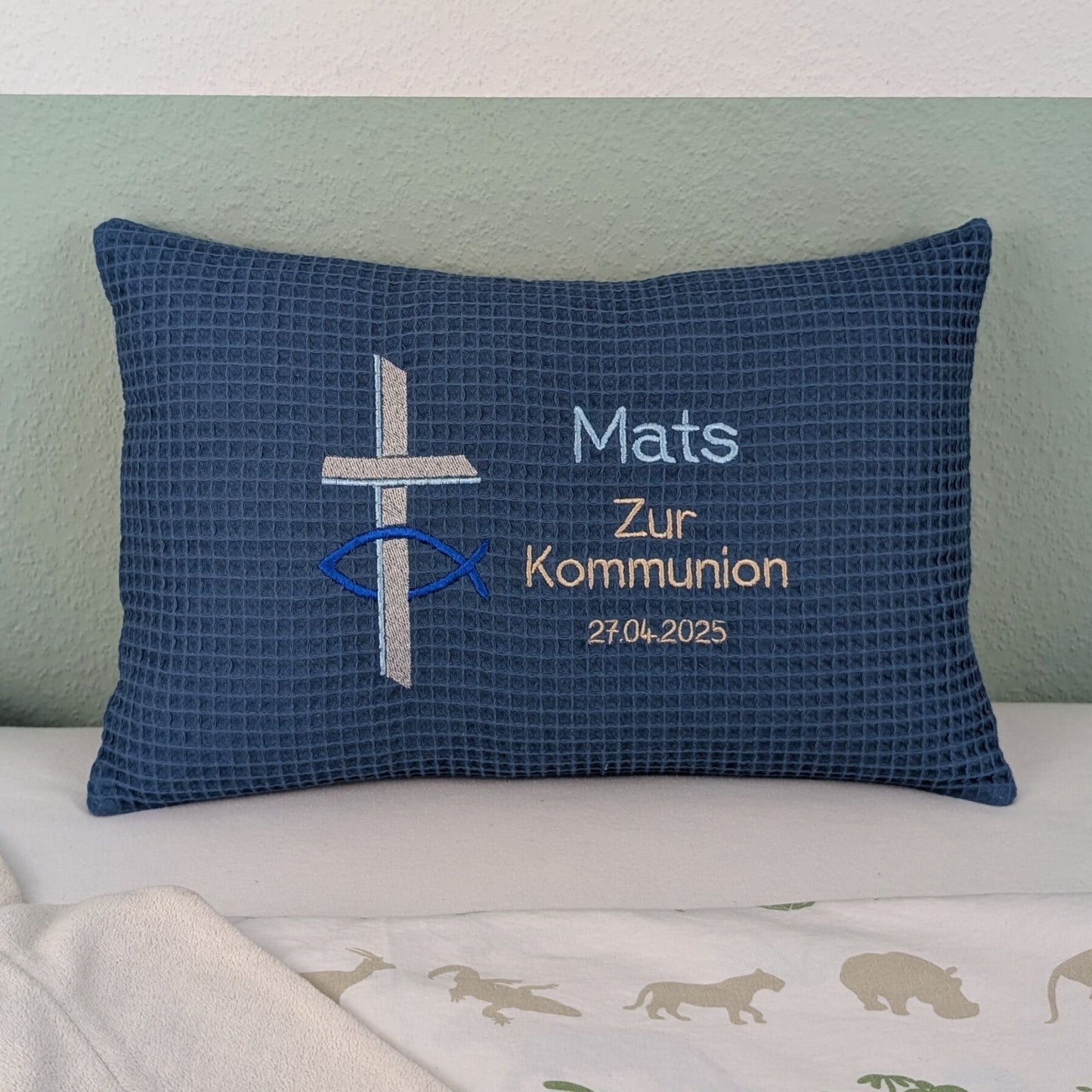 Rechteckiges Kissen in blauer Farbe mit Waffelstruktur liegt auf einem Bett. Auf dem Kissen sind ein gesticktes Kreuz und ein christliches Ichthys-Symbol (Fisch) sowie der Text „Mats – Zur Kommunion – 27.04.2025“ in verschiedenen Farben eingestickt. Das Kissen dient als personalisiertes Geschenk zur Erstkommunion.