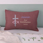 Rechteckiges Kissen in altrosa Farbe mit Waffelstruktur liegt auf einem Bett. Auf dem Kissen sind ein gesticktes Kreuz und ein christliches Ichthys-Symbol sowie der Text „Jasmin – Zur Kommunion – 11.5.2025“ in verschiedenen Farben eingestickt. Das Kissen dient als personalisiertes Geschenk zur Erstkommunion.