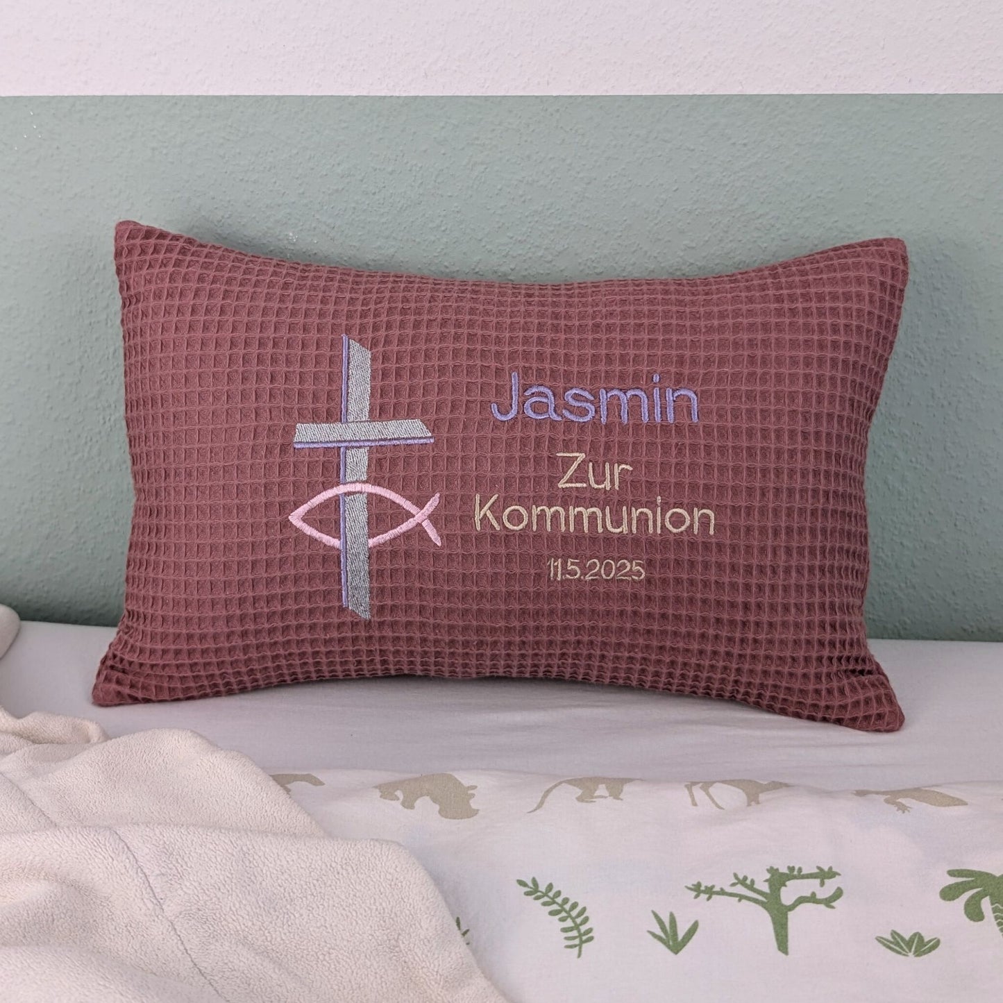 Rechteckiges Kissen in altrosa Farbe mit Waffelstruktur liegt auf einem Bett. Auf dem Kissen sind ein gesticktes Kreuz und ein christliches Ichthys-Symbol sowie der Text „Jasmin – Zur Kommunion – 11.5.2025“ in verschiedenen Farben eingestickt. Das Kissen dient als personalisiertes Geschenk zur Erstkommunion.