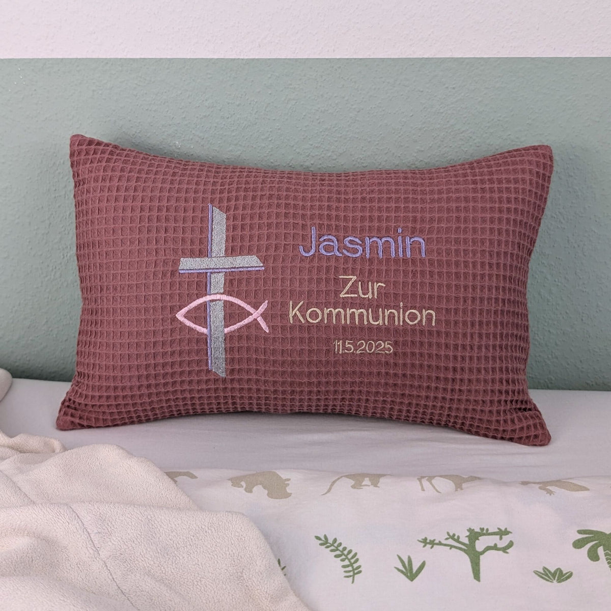 Rechteckiges Kissen in altrosa Farbe mit Waffelstruktur liegt auf einem Bett. Auf dem Kissen sind ein gesticktes Kreuz und ein christliches Ichthys-Symbol sowie der Text „Jasmin – Zur Kommunion – 11.5.2025“ in verschiedenen Farben eingestickt. Das Kissen dient als personalisiertes Geschenk zur Erstkommunion.