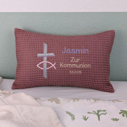 Rechteckiges Kissen in altrosa Farbe mit Waffelstruktur liegt auf einem Bett. Auf dem Kissen sind ein gesticktes Kreuz und ein christliches Ichthys-Symbol sowie der Text „Jasmin – Zur Kommunion – 11.5.2025“ in verschiedenen Farben eingestickt. Das Kissen dient als personalisiertes Geschenk zur Erstkommunion.