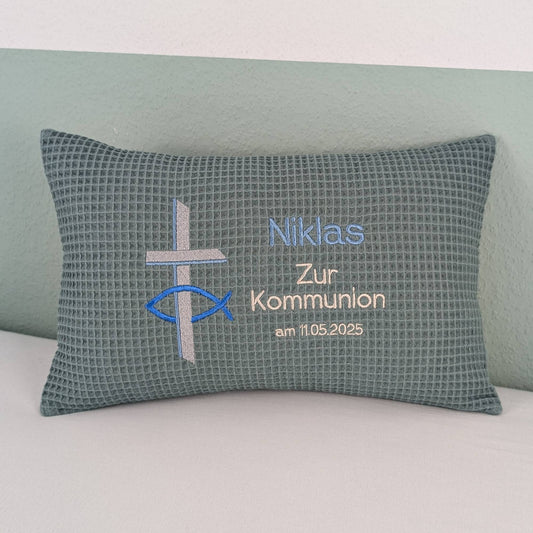 Rechteckiges Kissen in türkis Farbe mit Waffelstruktur liegt auf einem Bett. Auf dem Kissen sind ein gesticktes Kreuz und ein christliches Ichthys-Symbol (Fisch) sowie der Text „Niklas – Zur Kommunion – am 11.5.2025“ in Blau und silber Farben eingestickt. Das Kissen dient als personalisiertes Geschenk zur Erstkommunion.